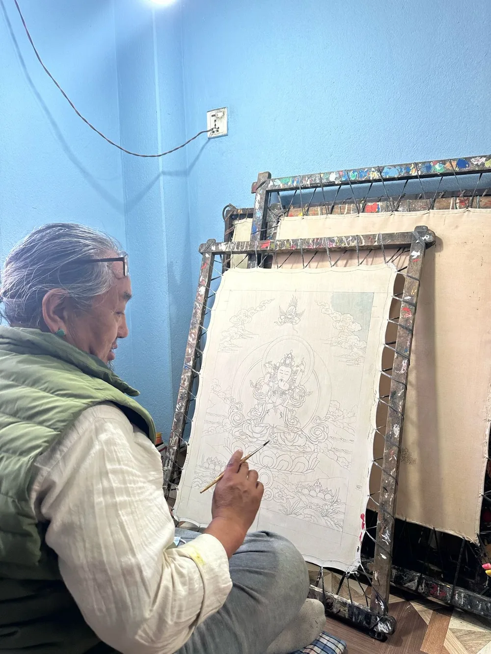 Master Tenzin Dhargay seated beside a thangka drawing mounted on a painting frame